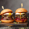 Two homemade beef burgers with mushrooms, micro greens, red onion, fried eggs and beet sauce on wooden cutting board. Side view, close up